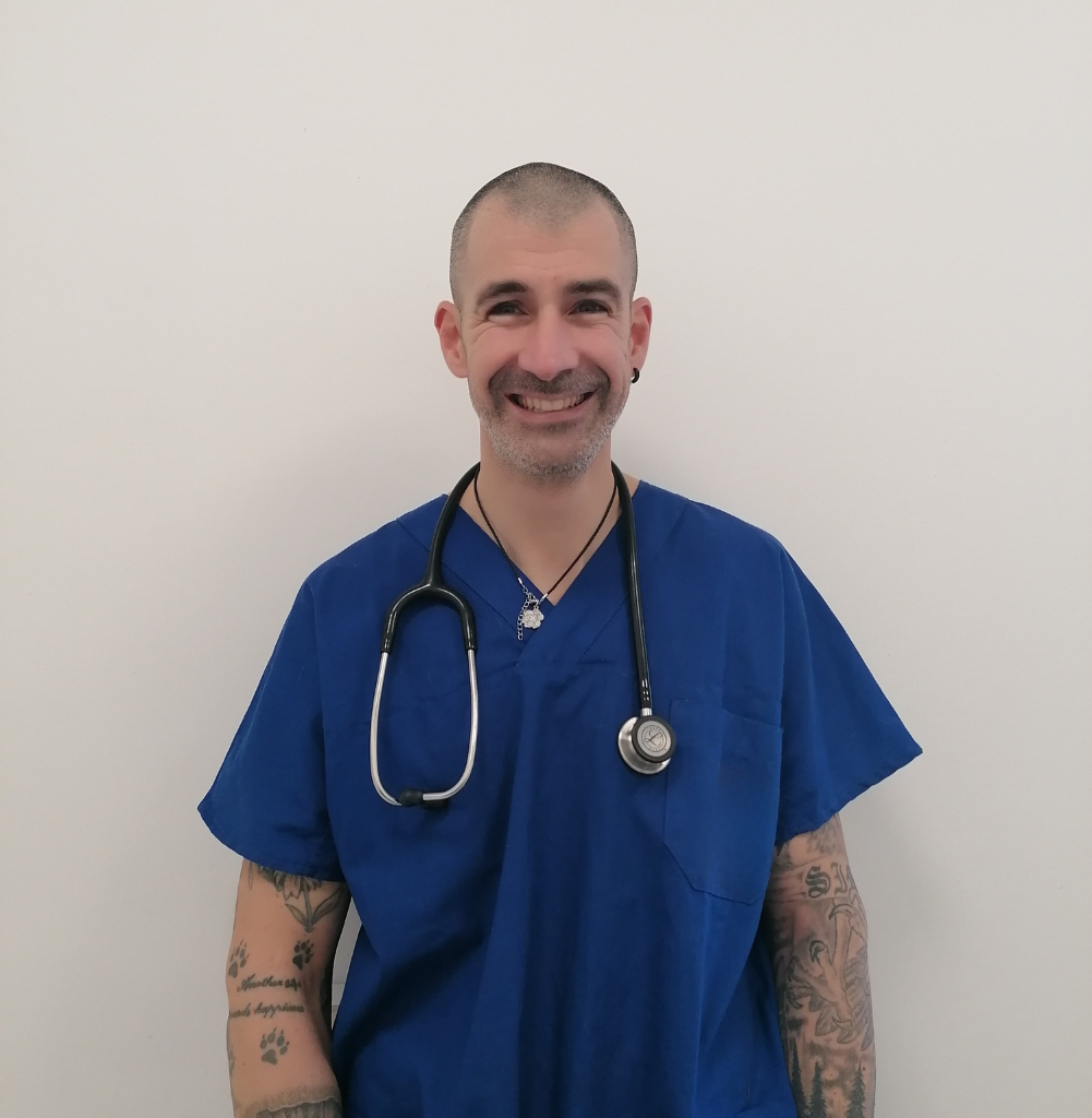 Un uomo sorridente che indossa un camice da medico blu e uno stetoscopio si staglia su un semplice sfondo bianco. Ha tatuaggi su entrambe le braccia e capelli corti, e rappresenta con orgoglio la sua squadra come veterinario a Vicenza.