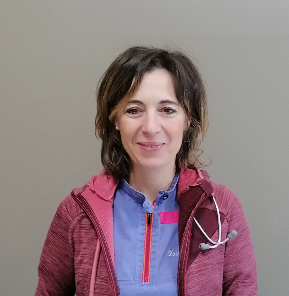 Una donna con i capelli castani lunghi fino alle spalle, che fa parte dell'équipe di Vetestudio a Schio, a Vicenza, sorride alla telecamera con un camice blu e una giacca con zip marrone. Uno stetoscopio è drappeggiato intorno al suo collo mentre si trova su uno sfondo semplice.