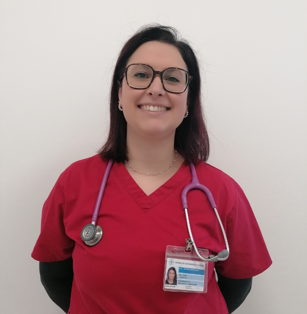 Una donna con un camice rosso e uno stetoscopio al collo sorride alla telecamera. Ha gli occhiali, i capelli scuri lunghi fino alle spalle, un tesserino di riconoscimento e fa parte di un'équipe dedicata del veterinario di Schio, Vicenza. Lo sfondo è semplice e bianco.