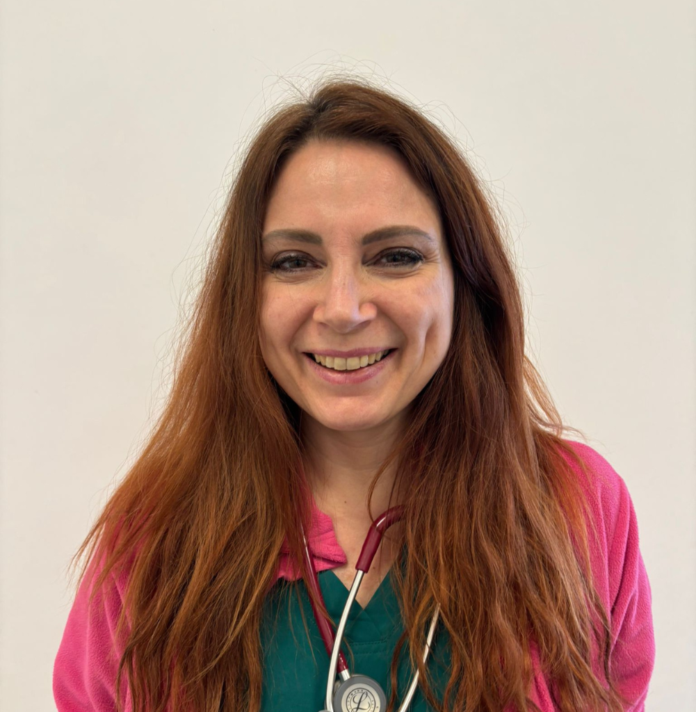 Una donna sorridente con lunghi capelli castano-rossicci, un top verde, una giacca rosa e uno stetoscopio al collo si staglia su uno sfondo bianco, parte dell'équipe di veterinari a Vicenza.