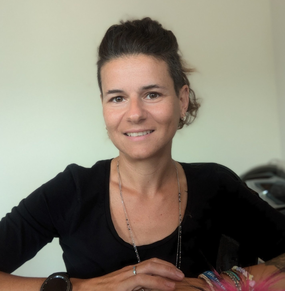 Una donna con i capelli corti e scuri, che indossa un top nero, una collana e un braccialetto, siede e sorride alla telecamera in un ambiente interno ben illuminato: un membro del team di Veterinario a Vicenza che si avvicina.