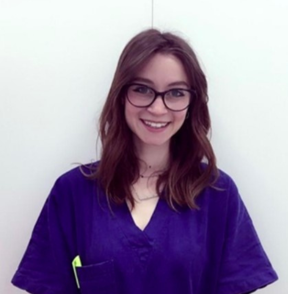 Una giovane donna con lunghi capelli castani e occhiali sorride all'obiettivo. Indossa un camice viola, su un semplice sfondo bianco, pronta a sostenere la sua squadra come veterinario a Schio.