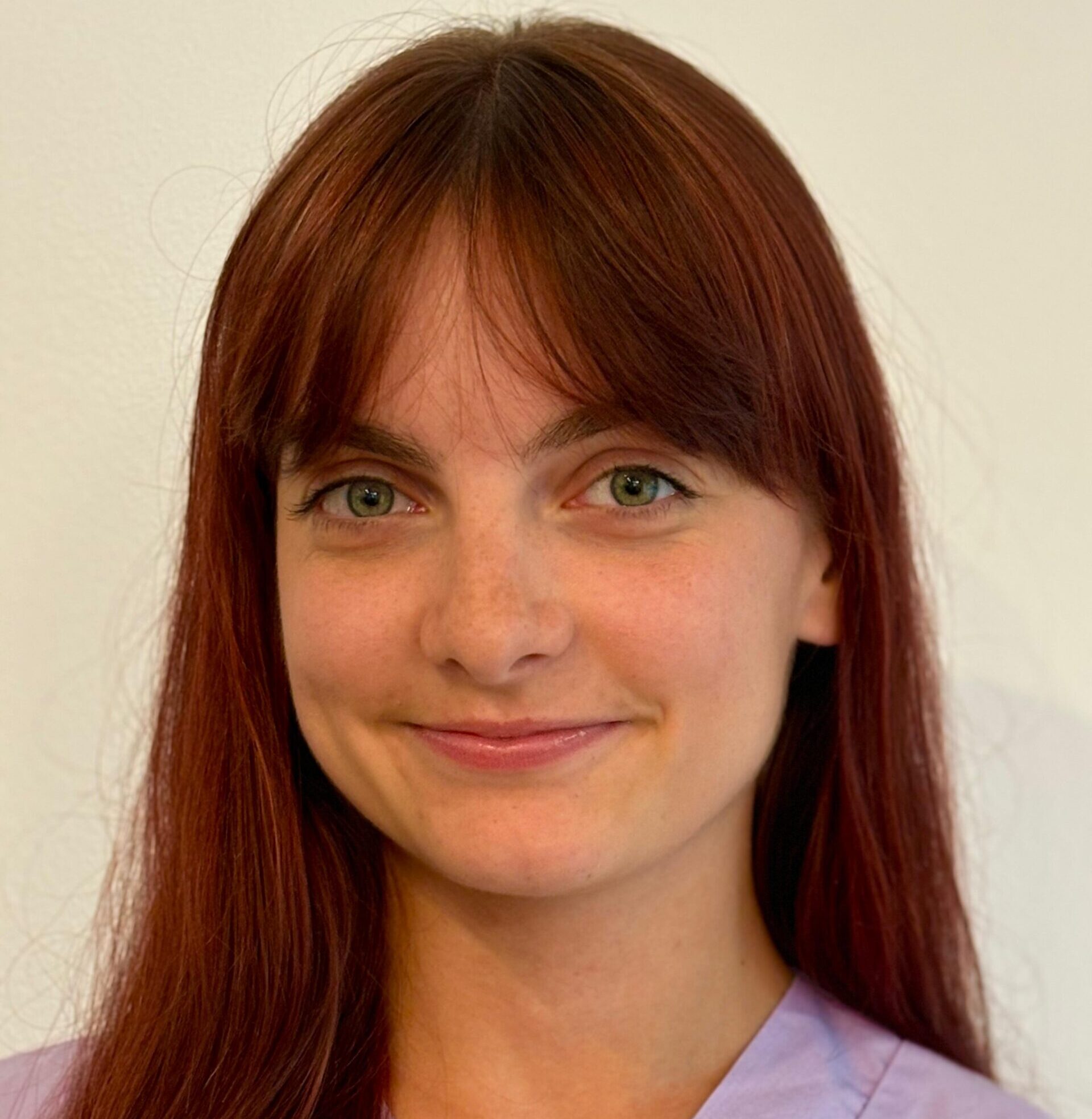 Una giovane donna con capelli lunghi e lisci color castano e frangetta sorride dolcemente. Indossa un top viola chiaro ed è in piedi davanti a uno sfondo chiaro, pronta a entrare nel team del veterinario di Vicenza.